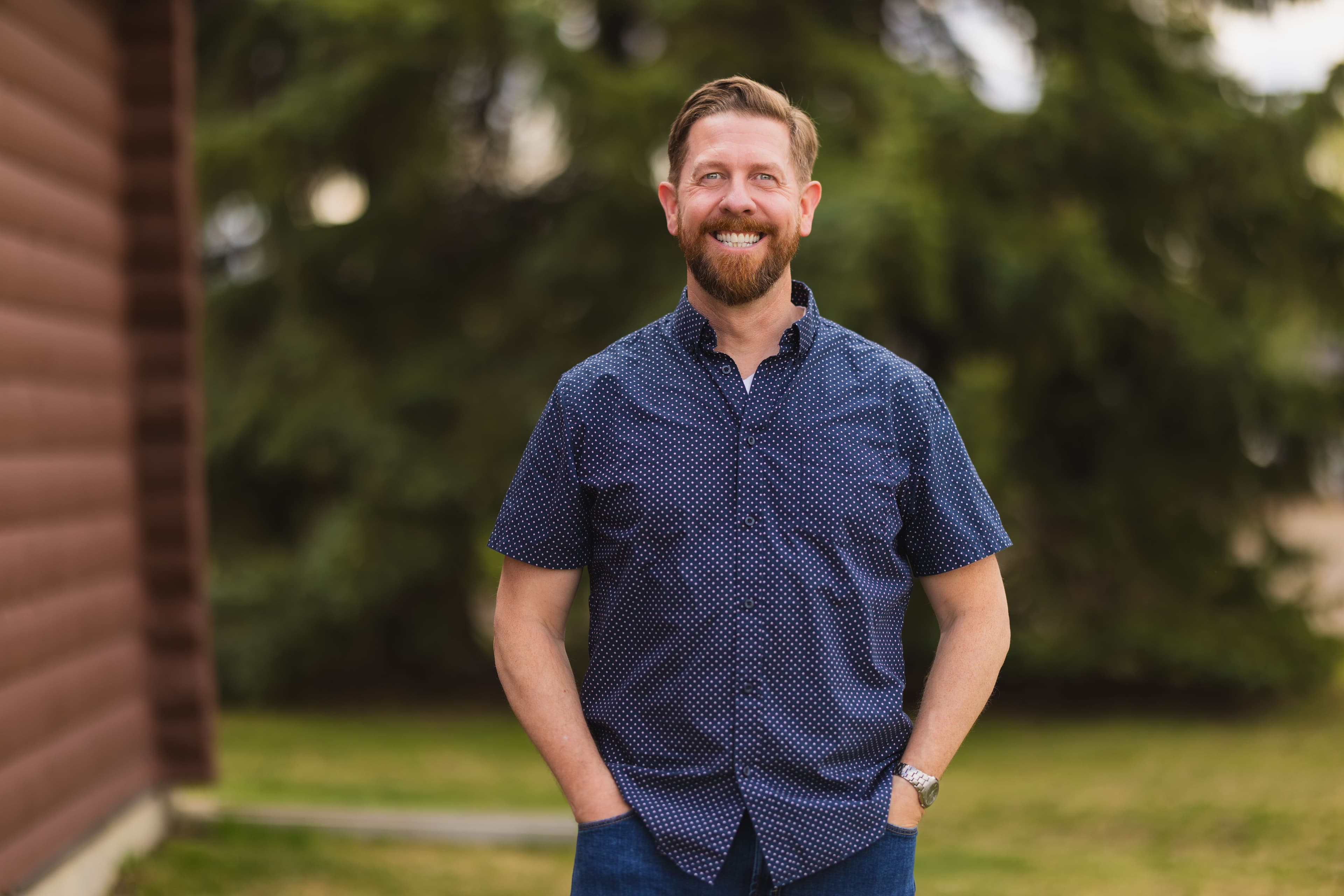 MLA Kyle Kasawski stands outside on green grass beside the Log Cabin in Sherwood Park.