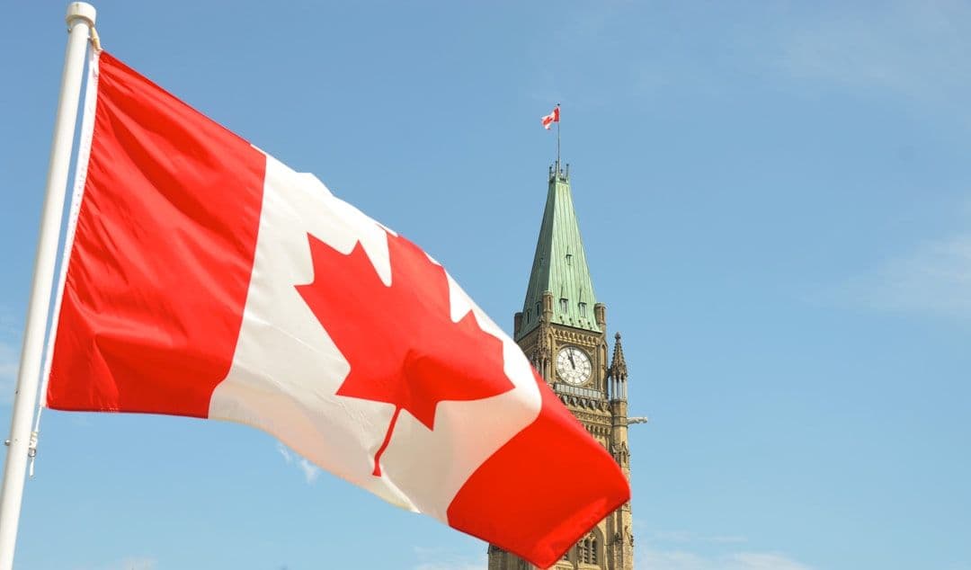 Canadian flag waves in the foreground, eclipsed by the House of Commons Peace Tower in Ottawa, Ontario.